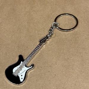 May include: A black and white electric guitar keychain with a silver key ring. The guitar has a white pickguard and silver strings. The keychain is made of metal and is a miniature replica of a guitar.