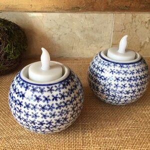 May include: Two white ceramic candle holders with blue floral patterns. Each holder has a white LED tea light inside.