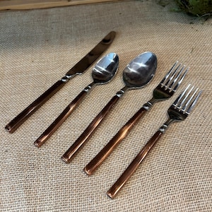 May include: A set of five pieces of silverware, including a knife, two spoons, and two forks. The silverware has a shiny silver finish on the eating surfaces and copper-colored handles. The pieces are arranged on a textured, tan surface.