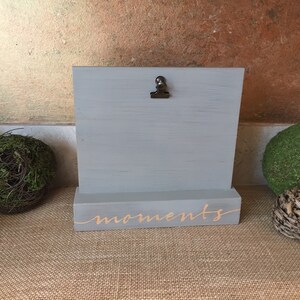 May include: Gray wooden picture frame with a metal clip and a base that says "moments" in gold lettering.