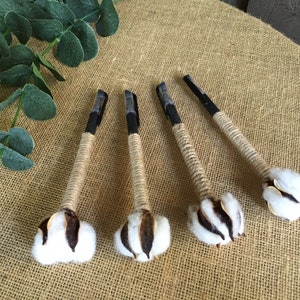 May include: Five black pens wrapped in twine with cotton bolls attached to the ends. The pens are lying on a burlap surface.