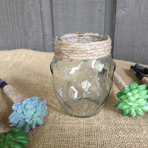 May include: A clear glass jar with a brown twine band around the top. The jar is sitting on a burlap surface with several small green succulent plants around it.