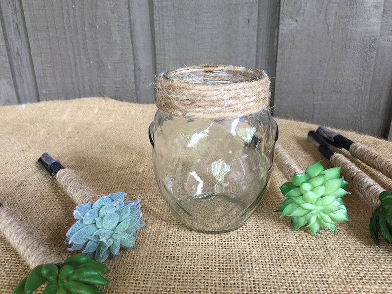 May include: A clear glass jar with a brown twine band around the top. The jar is sitting on a burlap surface with several small green succulent plants around it.