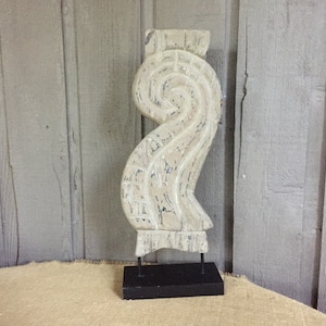 May include: A decorative, off-white wooden sculpture with a swirling design, mounted on a black metal stand. The sculpture has a distressed finish, revealing hints of blue paint. The piece is displayed on a neutral-toned surface.