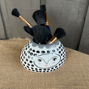May include: A white ceramic brush holder with black polka dots and a hand-painted face. The holder has a gray rim and is filled with black makeup brushes.