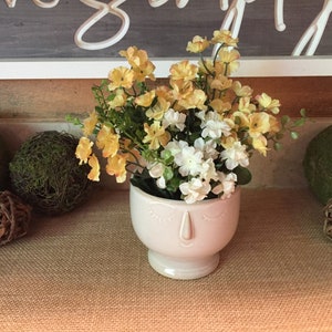 May include: A cream-colored ceramic planter shaped like a human face with closed eyes and a nose, filled with a bouquet of artificial yellow and white flowers with green stems and leaves, positioned centrally in the frame.