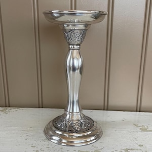 May include: Silver candle holder with an ornate design featuring grapes and leaves. The holder has a round base and a tall, slender stem.