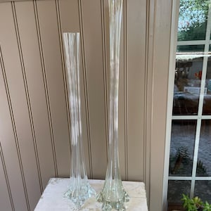 May include: Two tall, clear glass vases. The vases have a slender, fluted design that flares out at the base. The bases have a starburst pattern. The vases are displayed on a white, distressed wooden surface.