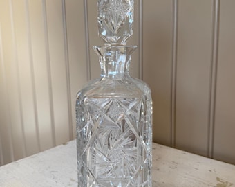 May include: A clear, cut-glass decanter with a square body and a matching stopper. The glass is intricately cut with starburst and geometric patterns. The decanter is designed to hold liquids and is a decorative item.