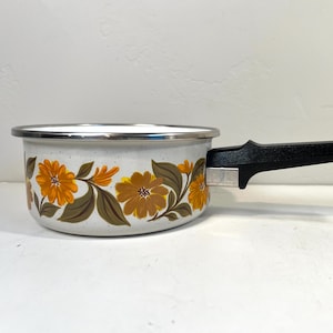 May include: A vintage-style saucepan with a white speckled finish and a floral pattern in shades of orange, brown, and yellow. It has a black handle and a silver rim. This retro kitchenware adds a touch of nostalgia to any kitchen.