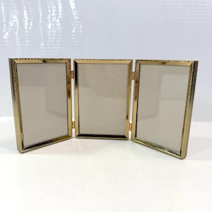 May include: A triptych photo frame featuring three vertical rectangular panels connected by metal hinges. The frame has a polished gold-tone finish with a textured, decorative pattern along the outer edges and includes light beige matting.