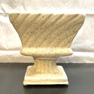 May include: Cream-colored ceramic planter with a square base and a pedestal. The planter has a flared, textured top with a spiral design and is speckled with brown flecks. The planter is suitable for small plants or decorative use.