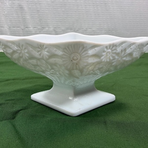 May include: A white milk glass bowl featuring a square base and a scalloped rim. The exterior surface is decorated with a raised relief pattern of daisies and leaves, with a cross-hatched texture near the base of the bowl.