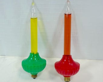 May include: Two bubble light bulbs with clear glass tubes filled with liquid. One bulb has a yellow liquid and a green base, while the other has an orange liquid and a red base. Both bulbs feature a standard metal screw-in base at the bottom.