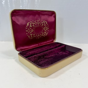 May include: A rectangular jewelry box with a cream-colored exterior and a hinged lid. The interior is lined with deep burgundy velvet, featuring a central slot. The inside of the lid displays a gold-colored ornate floral pattern on burgundy fabric.