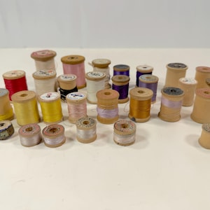 May include: A collection of vintage wooden sewing thread spools in various sizes and colors, including red, yellow, pink, purple, and white. Some spools are empty, while others are wound with thread, featuring paper labels on the ends.