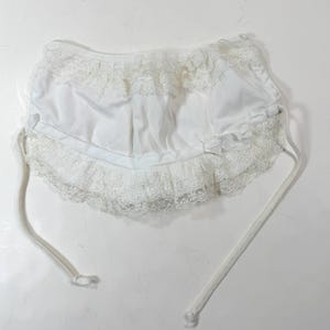 May include: White baby bonnet with lace trim and ties. The bonnet features a ruffled lace edge and a gathered crown. The ties are made of a soft, white cord. This vintage-style bonnet is perfect for a baby.