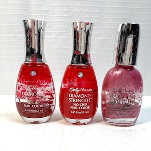 May include: Three bottles of Sally Hansen nail polish in shades of red and pink. The bottles have silver caps and are labeled "Diamond Strength" and "Maximum Growth Plus." Each bottle contains 0.45 fl oz / 13.3 ml of nail color.