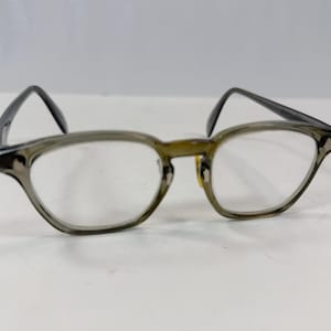 May include: Eyeglasses with a translucent olive-gray frame and clear lenses. The frame features a rounded rectangular shape with small silver-toned metal accents at the outer corners and a bridge connecting the two rims.