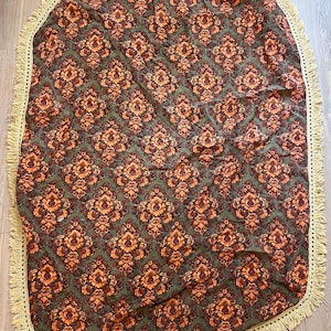 May include: Oval-shaped, patterned textile featuring a repeating floral design in shades of orange and brown on a green background. The edge is trimmed with a cream-colored fringe, adding a decorative element to the textile.