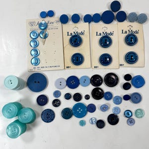 May include: A collection of blue buttons in various shades and sizes. Some buttons are on cards labeled "La Mode" and "Je Tone". The buttons are made of different materials and have varying designs, including four-hole and two-hole styles.