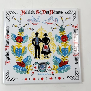 May include: Square white tile with a folk art design. Features a couple in traditional clothing, floral and scroll motifs in red, blue, and yellow, and text in Swedish. The tile has a glossy finish.