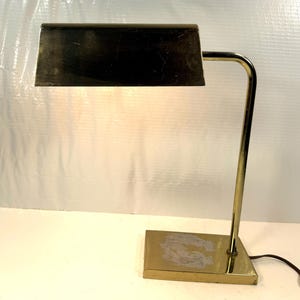 May include: A vintage brass desk lamp with a rectangular shade and a curved arm. The lamp has a rectangular base and a cord for power. The shade is dark, and the lamp is lit, casting a warm glow.
