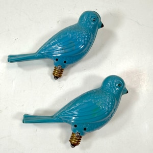 May include: Two bright blue bird-shaped light bulbs with molded feather textures, black painted eyes, and small black beaks. Each bird figure features a standard brass-colored metal screw base extending from the underside of the body.