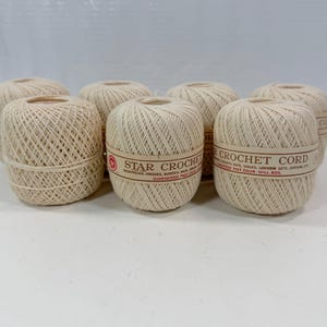 May include: Seven balls of off-white Star Crochet Cord, each wrapped in a paper band featuring red text and a star logo. The yarn is wound into a compact, rounded shape, displaying a consistent, textured weave throughout the entire surface.