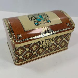 May include: A decorative metal chest with a domed lid. The chest features a brown and gold color scheme with a repeating fleur-de-lis pattern. The lid has a crest design with teal accents. The edges are trimmed with gold beading.