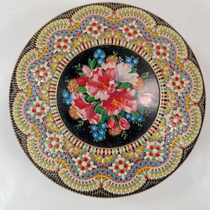 May include: A round metal tin featuring a central black circular panel decorated with a vibrant bouquet of pink and white flowers, surrounded by an intricate, colorful border of repeating floral patterns in red, yellow, green, and white.