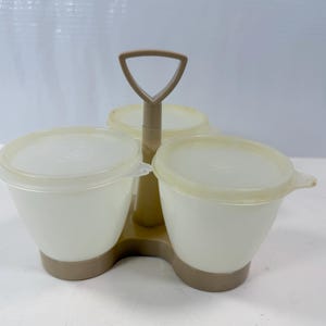 May include: A vintage three-part serving dish with a handle. The set includes three white, lidded bowls with a light brown base and handle. The lids are translucent. The serving dish is designed to hold condiments or snacks.