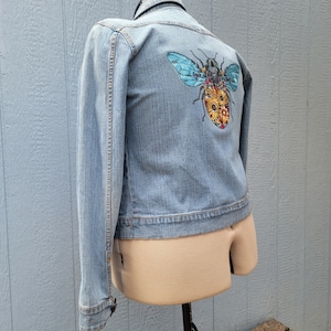 May include: A light blue denim jacket with a colorful embroidered bee on the back.