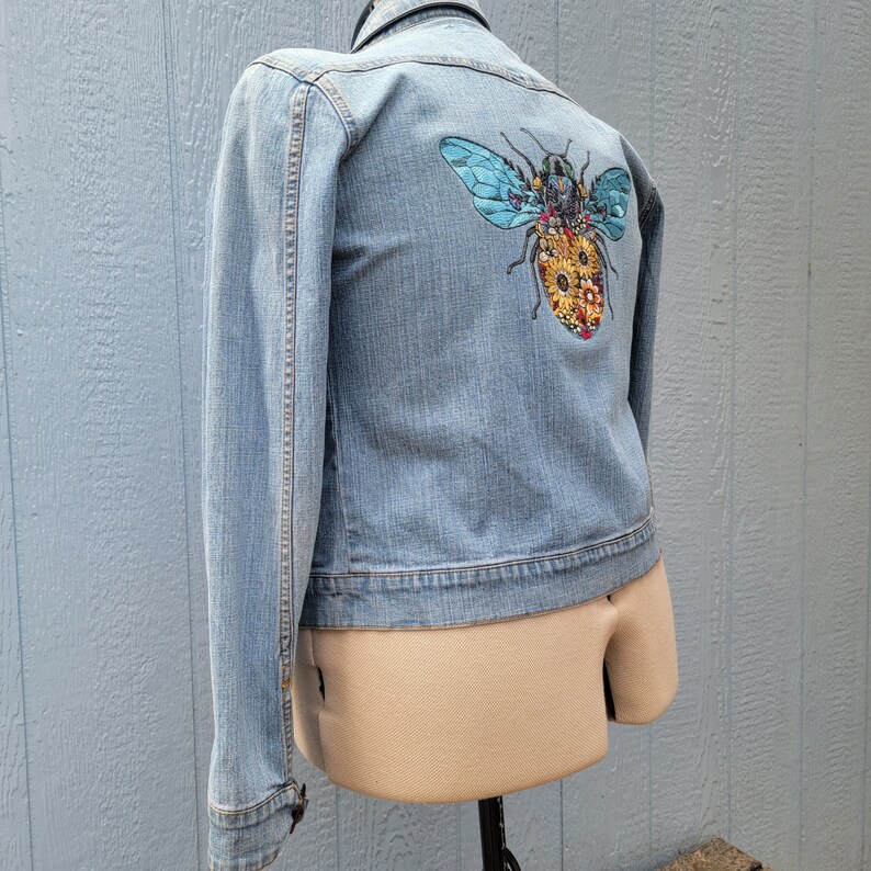 May include: A light blue denim jacket with a colorful embroidered bee on the back.