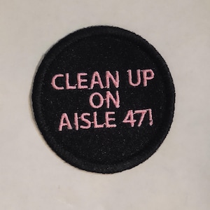 May include: A round, black embroidered patch with the phrase "CLEAN UP ON AISLE 47!" in pink thread. The patch has a textured appearance and is likely designed to be sewn or ironed onto fabric.