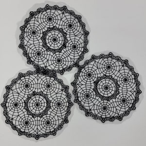 May include: Three black lace doilies with a spiderweb design. The doilies are circular and have a delicate, intricate pattern.