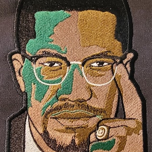 May include: Embroidered patch of a man with a green and gold face, wearing glasses and a suit. He is holding a cigarette in his right hand.