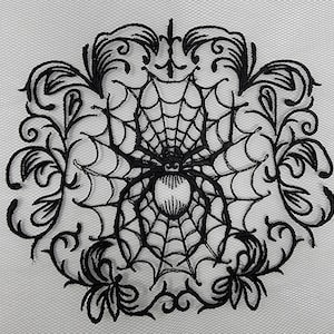May include: Black embroidered design of a spider in a web with intricate floral details. The design is on a white background.