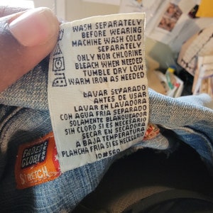 May include: A white care label with instructions for washing and ironing a garment. The label has text in both English and Spanish. The text reads "Wash separately before wearing. Machine wash cold separately. Only non chlorine bleach when needed. Tumble dry low. Warm iron as needed. Lavar separado antes de usar. Lavar en lavadora con agua fria separado. Solamente blanqueador sin cloro si es necesario. Secar en secadora a baja temperatura. Plancha fria si es necesario. 10#560"