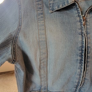 May include: A close-up of a light blue denim jacket with a zipper closure. The jacket has a seam down the center of the back and a seam on each side of the front.
