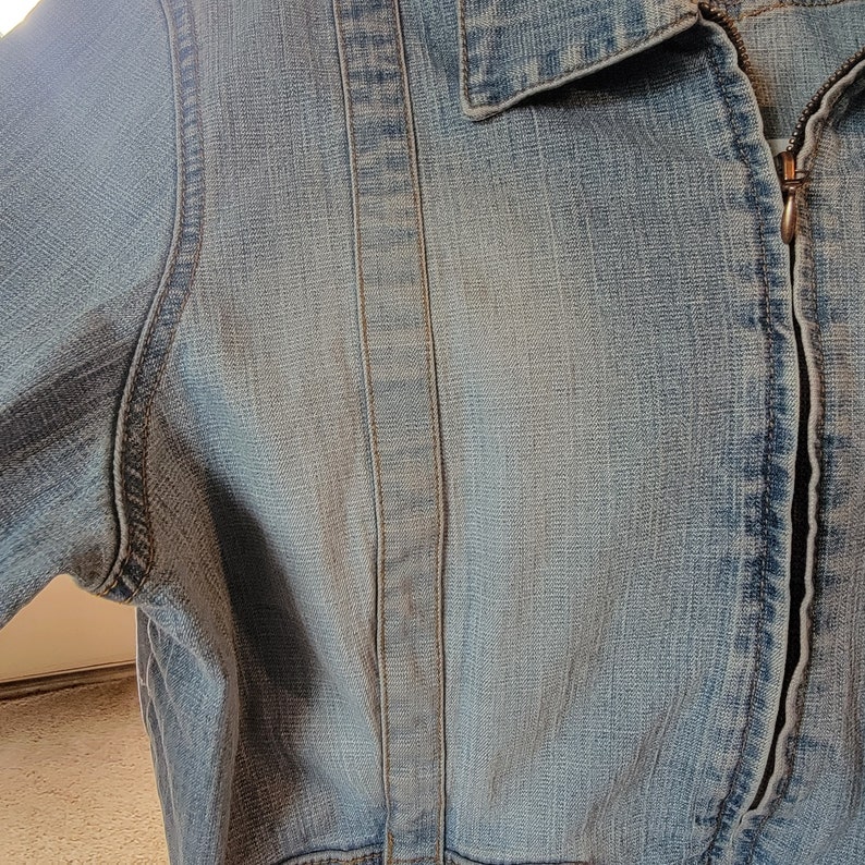 May include: A close-up of a light blue denim jacket with a zipper closure. The jacket has a seam down the center of the back and a seam on each side of the front.