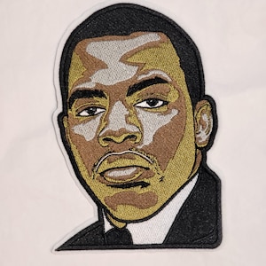 May include: An embroidered patch featuring a portrait of a person with short dark hair, wearing a black suit jacket and a white collared shirt. The design uses shades of brown, gold, and silver thread to create highlights and shadows on the face.