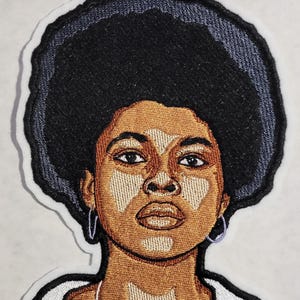 May include: An embroidered patch featuring a portrait of a person with a large black afro hairstyle, brown skin with lighter patches, and small hoop earrings. The design is outlined in black thread with a white border against a plain background.