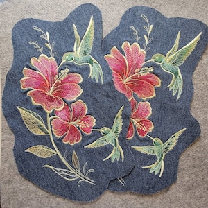 May include: Two denim patches with embroidered images of pink hibiscus flowers and green hummingbirds. The patches are on a gray felt background.