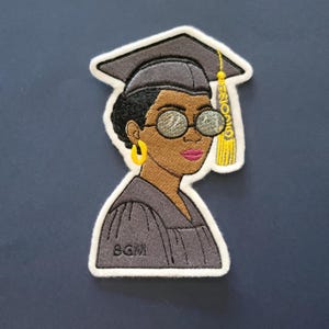 May include: Embroidered patch featuring a Black woman in a graduation cap and gown. The patch has a white border and includes the text "2020" on the tassel. The woman wears glasses and yellow hoop earrings. The initials "SGM" are on the gown.