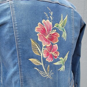May include: A denim jacket with embroidered flowers and hummingbirds. The flowers are pink and red with gold accents. The hummingbirds are gold and white.