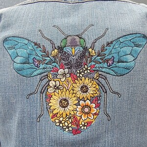 May include: Embroidered denim jacket with a blue and yellow bee design. The bee is surrounded by colorful flowers and has a green head.