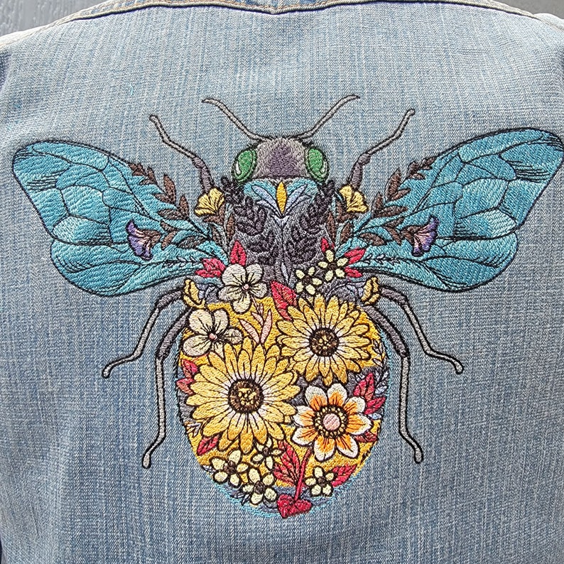 May include: Embroidered denim jacket with a blue and yellow bee design. The bee is surrounded by colorful flowers and has a green head.