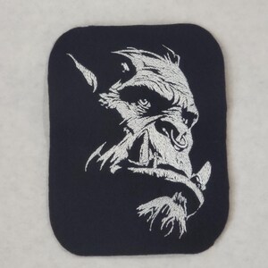 May include: A rectangular black fabric patch featuring a white embroidered graphic of an orc face in profile. The design highlights the creature's prominent tusks, a nose ring, and a textured beard, rendered in detailed white thread against the dark background.