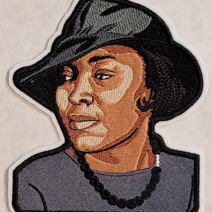 May include: An embroidered patch featuring a portrait of a person wearing a black wide-brimmed hat and a dark gray shirt. The person has brown skin with lighter patches, dark eyes, and wears a black beaded necklace. The patch has a white border.